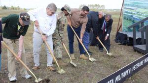 Five people stick shovels into the ground, while a sixth person assists one of them.
